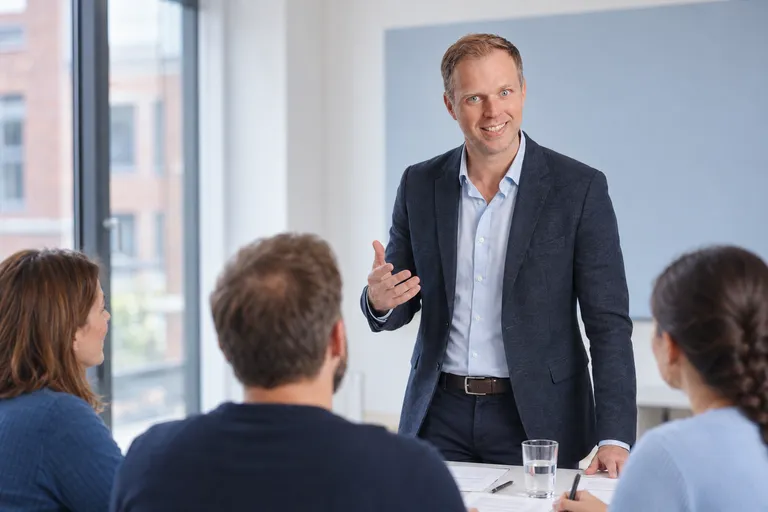 Dennis Tefett spricht nah an den Tischen mit drei Teilnehmenden, hinter ihm die blaue Tafel und das Fenster des Seminarraums.